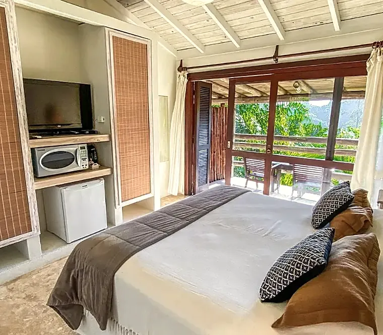 Quarto da casa com cama arrumada e porta aberta para a varanda com vista para a mata.