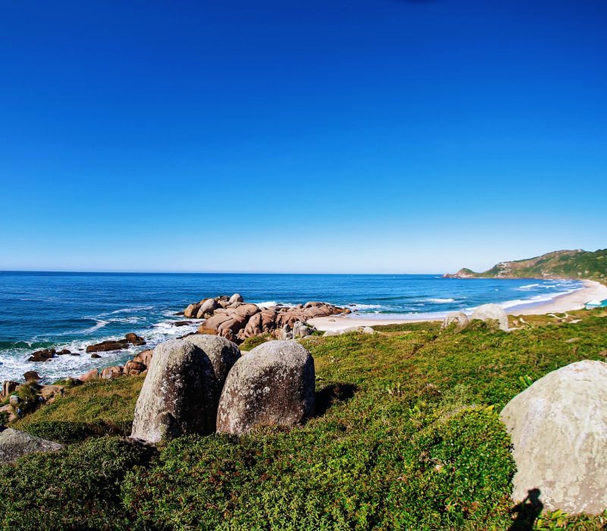 Costão de pedras da Praia do Rosa, com o mar aberto e a mata do morro ao fundo.